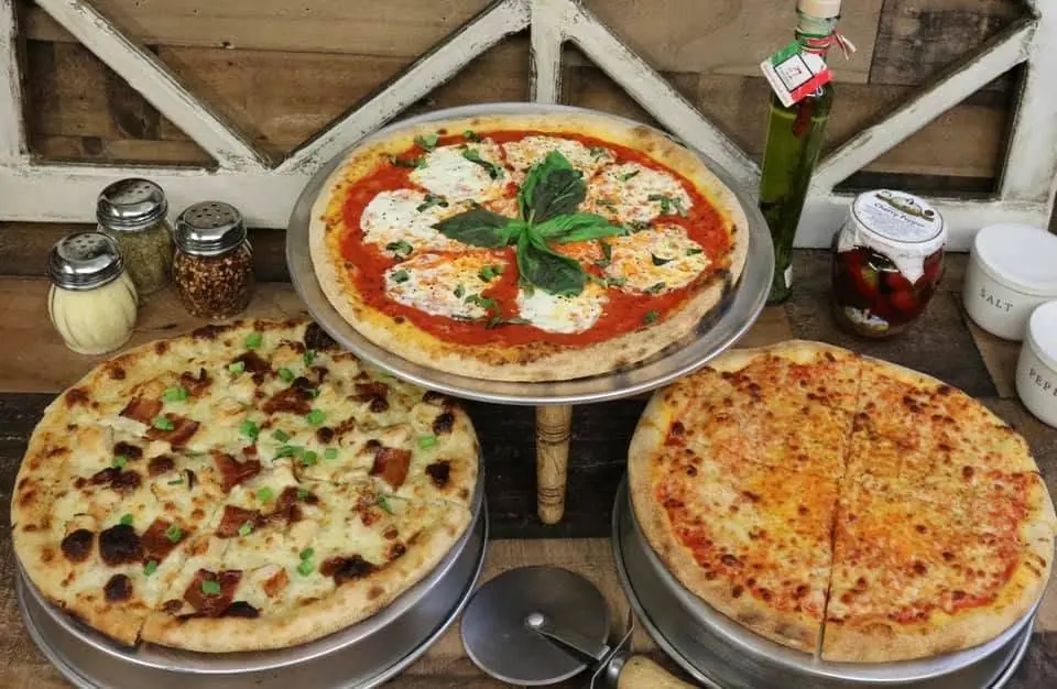 Three pizzas on trays on a wood counter, including a basil-topped margherita